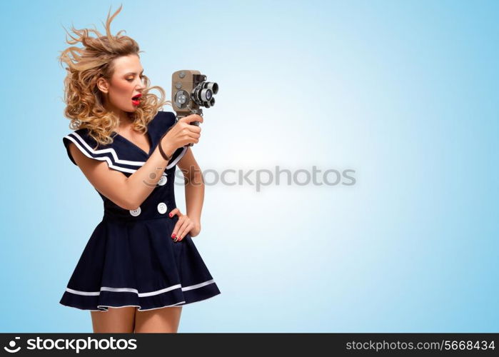 Retro photo of a glamorous pin-up sailor girl with an old vintage cinema 8 mm camera shooting a movie on blue background.