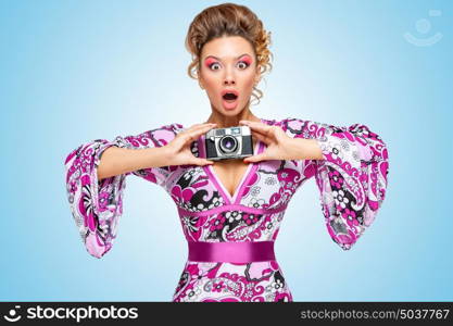 Retro photo of an amazed fashionable hippie homemaker, holding an old vintage photo camera with two hands and showing emotions on blue background.