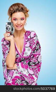 Retro photo of an amazed fashionable hippie homemaker with an old vintage photo camera showing emotions on blue background.