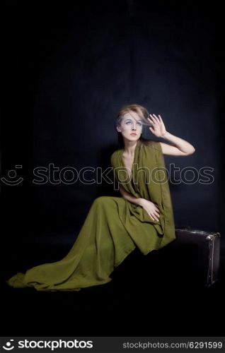 Retro portrait of beautiful young woman in long green dress, sitting on old suitcase