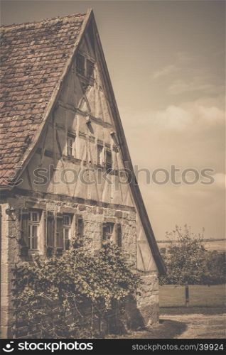 Retro style image of an antique german house with traditional architecture, gable roof, half timbered wall for the loft and stone wall, wooden shutters and vine bushes for the ground floor.