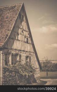 Retro style image of an antique german house with traditional architecture, gable roof, half timbered wall for the loft and stone wall, wooden shutters and vine bushes for the ground floor.