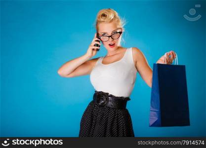 Retro style. Young woman pinup girl in glasses with shopping bag talking on cell phone. Blue background.