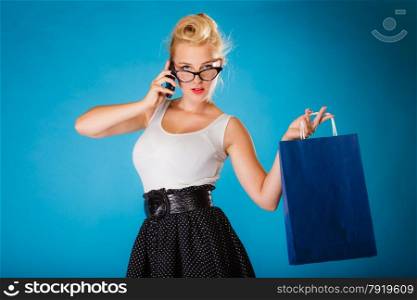 Retro style. Young woman pinup girl in glasses with shopping bag talking on cell phone. Blue background.
