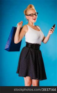 Retro style. Young woman pinup girl in glasses with shopping bag using on cell phone. Blue background.