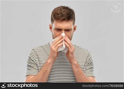 rhinitis, cold and allergy concept - young man with paper tissue blowing his nose over gray background. young man with paper tissue blowing his nose