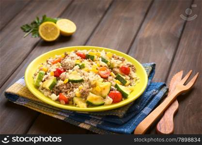 Rice dish with mincemeat and vegetables (sweet corn, cherry tomato, zucchini, onion) served on plate with wooden cutlery on the side, photographed on dark wood with natural light (Selective Focus, Focus in the middle of the dish)