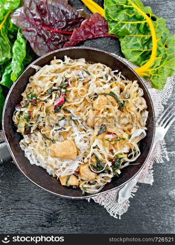 Rice noodles with leafy beet, chicken breast meat, cashew nuts and soy sauce in a frying pan on burlap on the background of black wooden board from above