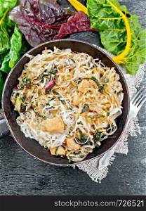 Rice noodles with leafy beet, chicken breast meat, cashew nuts and soy sauce in a frying pan on burlap on the background of black wooden board from above