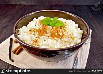 Rice porridge with cinnamon and mint in a brown bowl, sticks, spoon, vanilla pod on a napkin on a wooden board background