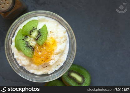 Rice pudding with kiwi pieces, orange jam and cinnamon in small glass bowl, photographed overhead on slate with natural light