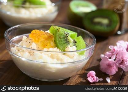 Rice pudding with kiwi pieces, orange jam and cinnamon in small glass bowl, photographed on dark wood with natural light (Selective Focus, Focus on the kiwi piece in the front)