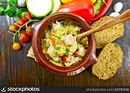Rice with chicken, bell pepper, zucchini and carrots in a clay pot with a spoon on napkin on dark wooden board background from above