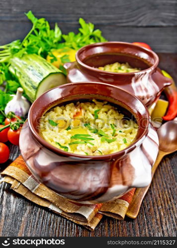 Rice with chicken, bell pepper, zucchini and carrots in two clay pots on a napkin, spoon and vegetables on dark wooden board background