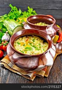 Rice with chicken, bell pepper, zucchini and carrots in two clay pots on a napkin, spoon and vegetables on dark wooden board background