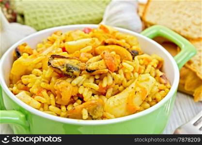 Rice with seafood and saffron in a green pot, napkin, fork, garlic bread on a wooden boards background