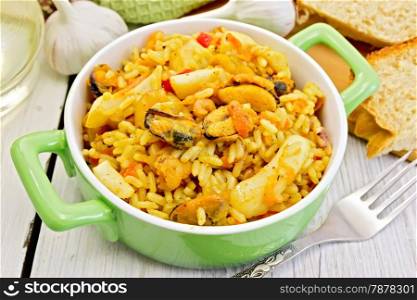 Rice with seafood and saffron in a green pot, napkin, fork, garlic bread on the background light wooden boards