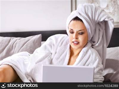 Rich woman in white bathrobe on a bed with laptop