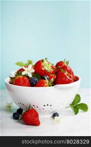 Ripe berries with leaves in white colander on marble table on blurred background
