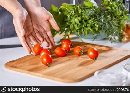 Ripe cherry tomatoes lay on the wooden board of the woman&rsquo;s hands on the kitchen table with fresh greens. Salad preparation. Space for the text. Woman&rsquo;s hands are putting ripe cherry tomatoes on a wooden board on the kitchen table with parsley. Copy space for text