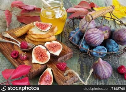 Ripe figs and blue plums in a vintage metal basket with wooden handle, sweet golden honey in a glass jar, raspberries and toast as well as red autumn leaves are on the old wooden table