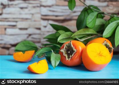 Ripe persimmon with slices on a blue table against a background of green leaves. Ripe persimmon with slices on blue table against green leaves