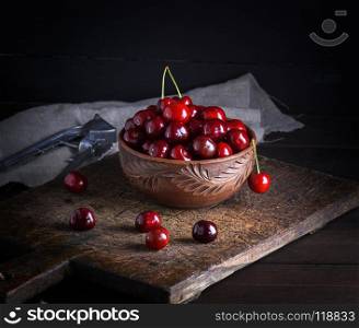ripe red cherry in a round clay bowl on a brown wooden board, black background. ripe red cherry in a round clay bowl
