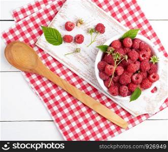 ripe red raspberries in a white wooden plate on a table of boards, next is a spoon and a textile towel, top view