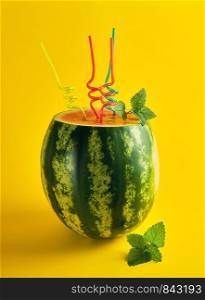 ripe striped green watermelon is cut and multicolored plastic cocktail tubes with a sprig of mint on a yellow background, refreshing summer cocktail for a friendly company