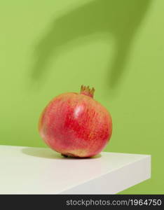 ripe, whole red pomegranate lies on a white table, shadow from the hand reaches for the fruit. Green background