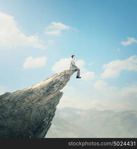 Risk in business. Young businessman sitting on edge of rock mountain