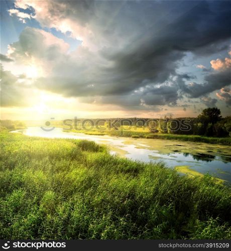 River flows through the field with lush green grass under a dramatic sky. River flows through the field with lush green grass