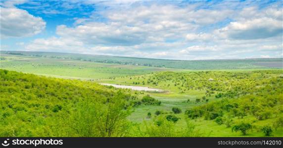 River valley with hills and terraces. On the slopes grow shrubs, trees and grass. In the blue sky, light cumulus clouds. Wide photo.
