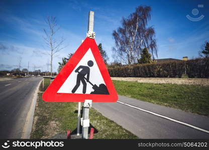 Road construction sign by a roadside in a city on a sunny day with blue sky