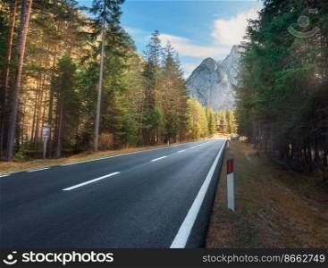 Road in autumn forest at sunset in Dolomites, Italy. Beautiful mountain roadway, orange tress, grass, high rocks, blue sky. Landscape with empty highway through the woods in fall. Travel background
