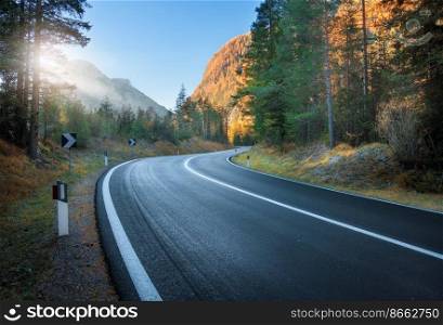 Road in autumn forest at sunset in Dolomites, Italy. Beautiful mountain roadway, orange tress, grass, high rocks, blue sky, sun. Landscape with empty highway through the woods in fall. Transportation