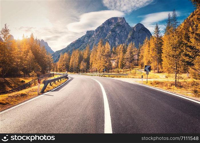 Road in autumn forest at sunset in Italy. Beautiful mountain roadway, trees with orange foliage and sunlight. Landscape with empty asphalt road through woodland, blue sky, high rocks in fall. Travel