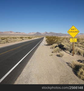 Road in desert with sign for soft shoulder and mountains in distance.