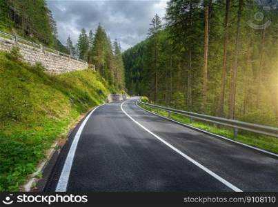 Road in green forest at sunset in summer. Dolomites, Italy. Beautiful mountain roadway, tress, grass, high rocks, blue sky with clouds. Landscape with empty highway through the wood in spring. Travel