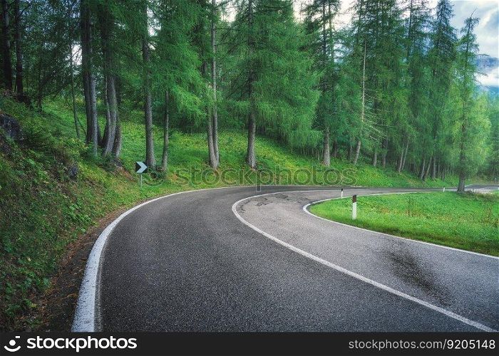 Road in green forest in rainy summer day. Dolomites, Italy. Beautiful mountain roadway, tress, grass, high rocks, blue sky with clouds. Landscape with empty highway through the wood in spring. Travel 