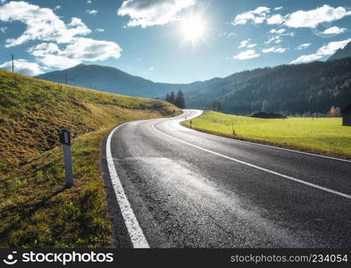 Road in mountain valley at sunny morning in Dolomites, Italy. View with asphalt roadway, meadows with green grass, mountains, blue sky with clouds and sun. Highway in fields. Trip in europe. Travel