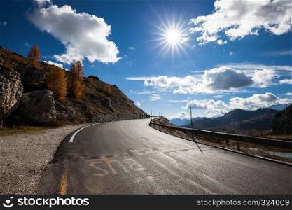 Road in mountain valley at sunny morning in Dolomites, Italy. View with asphalt roadway, meadows, mountains, blue sky with clouds and sun. Highway in mountain. Trip in europe. Travel. Road in mountain valley at sunny morning in Dolomites, Italy. View with asphalt roadway, meadows, mountains, blue sky with clouds and sun.