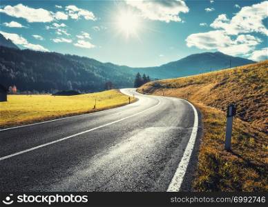 Road in mountain valley at sunny morning in Dolomites, Italy. View with asphalt roadway, meadows with green grass, mountains, blue sky with clouds and sun. Highway in fields. Trip in europe. Travel