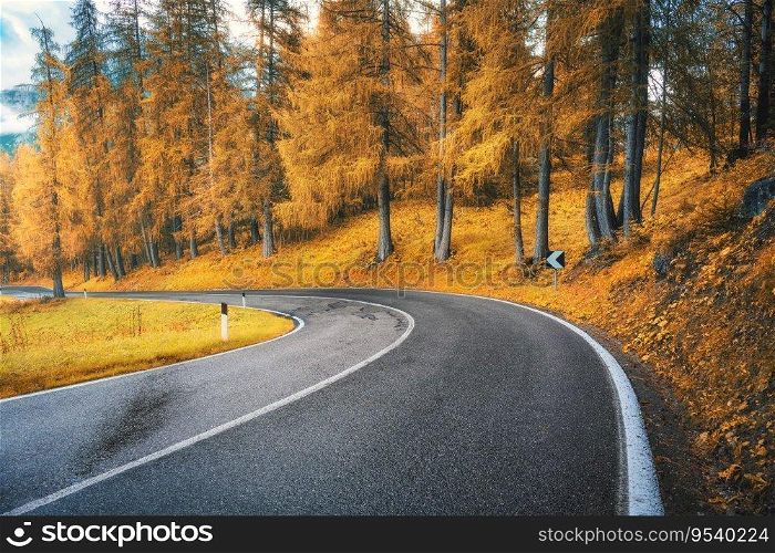 Road in orange forest at sunset in golden autumn. Dolomites, Italy. Beautiful mountain roadway, tress, grass, high rocks, blue sky with clouds. Landscape with empty highway through the woods in fall