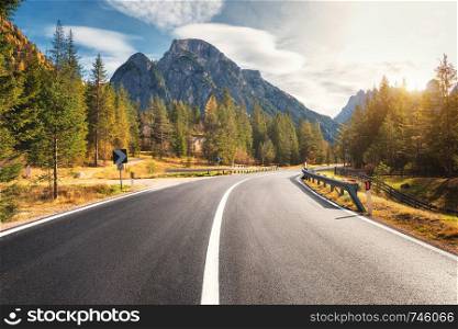 Road in summer forest at sunset in Italy. Beautiful mountain roadway, trees with green foliage and sunlight. Landscape with empty asphalt road through woodland, blue sky, high rocks. Travel in Europe