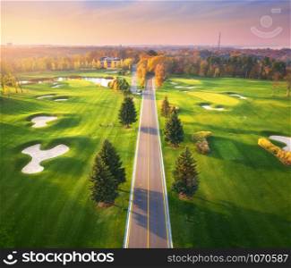 Road through the golf course at sunset in autumn. Aerial view of roadway and green golf field in fall. Landscape with route, meadow, green pine trees, orange forest, beautiful sky. View from above