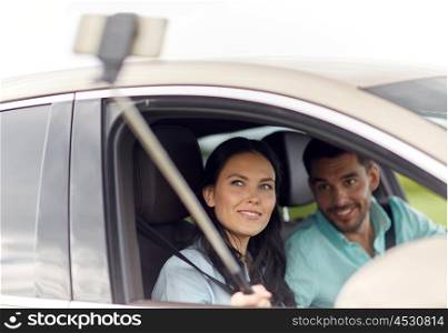 road trip, leisure, couple, technology and people concept - happy man and woman driving in car and taking picture with smartphone selfie stick