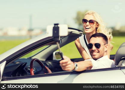 road trip, leisure, couple, technology and people concept - happy man and woman driving in cabriolet car and taking picture with smartphone on selfie stick