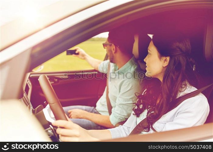 road trip, leisure, couple, technology and people concept - happy man and woman driving in car and taking selfie with smartphone. happy couple in car taking selfie with smartphone