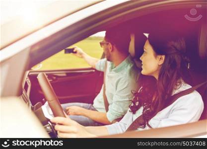 road trip, leisure, couple, technology and people concept - happy man and woman driving in car and taking selfie with smartphone. happy couple in car taking selfie with smartphone
